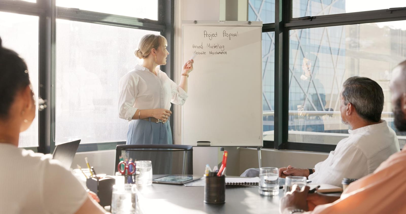 Mulher escreve tópicos em um flipchart diante de colegas sentados à mesa de reunião com vista para prédios.