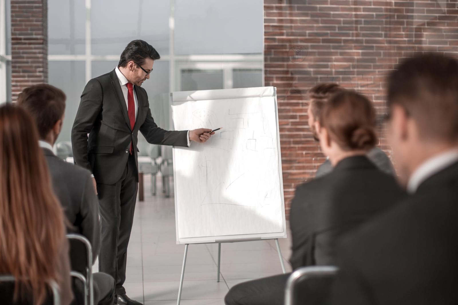 Homem de terno aponta para um flipchart diante de uma plateia pequena em uma sala com parede de tijolos.