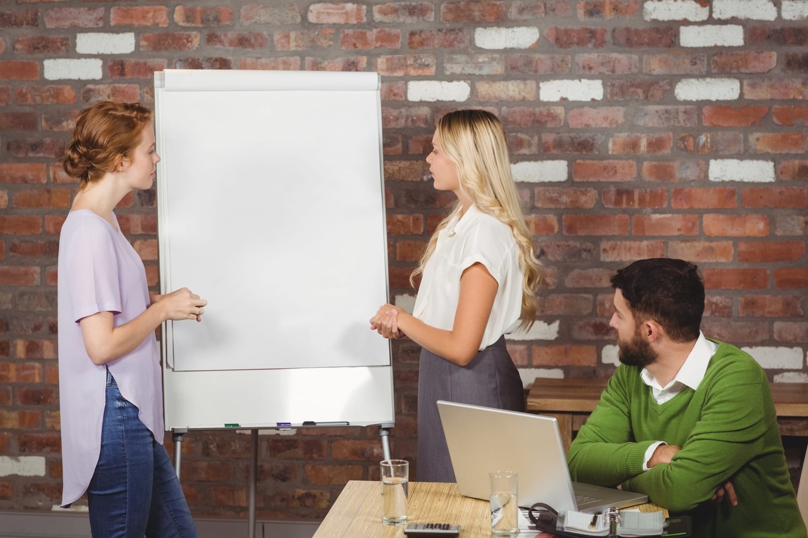 Duas mulheres conversam ao lado de um flipchart enquanto um colega acompanha sentado com notebook.